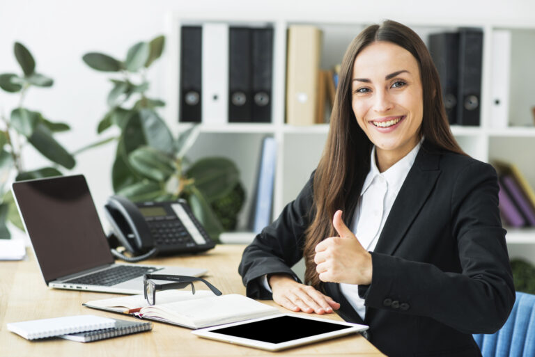 smiling-young-businesswoman-sitting-workplace-showing-thumb-up-sign